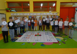 Grupa dzieci stoi pod tablicą, w ręku trzymają zegary papierowe.
